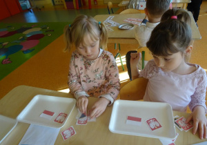 Dwie dziewczynki siedzą przy stoliku, na którym na tackach mają ułożone godło i mapę Polski.